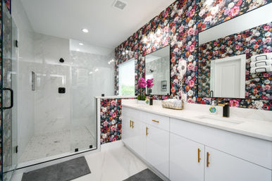 Contemporary master bathroom with bold floral wallpaper, glass-enclosed marble walk-in shower, white double-sink vanity with gold handles, bright mirrors, pink orchids on the quartz countertop and gray floor mats.