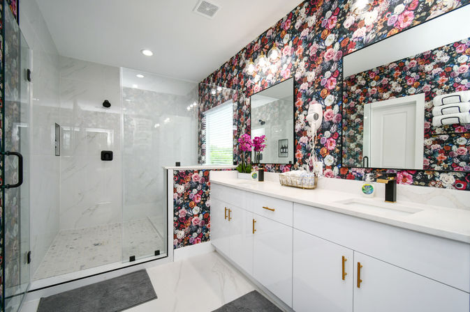 Contemporary master bathroom with bold floral wallpaper, glass-enclosed marble walk-in shower, white double-sink vanity with gold handles, bright mirrors, pink orchids on the quartz countertop and gray floor mats.