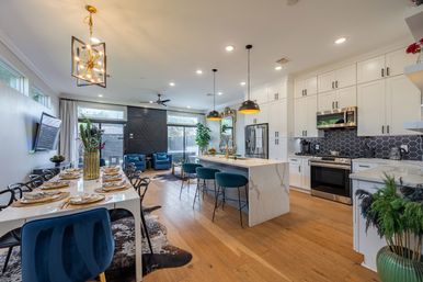 Contemporary open-plan kitchen and dining area with white shaker cabinets, marble waterfall island, black hex tile backsplash, brass pendant lights, teal bar stools, velvet dining chairs, hardwood floors and large glass sliding door.