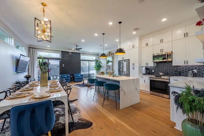 Contemporary open-plan kitchen and dining area with white shaker cabinets, marble waterfall island, black hex tile backsplash, brass pendant lights, teal bar stools, velvet dining chairs, hardwood floors and large glass sliding door.