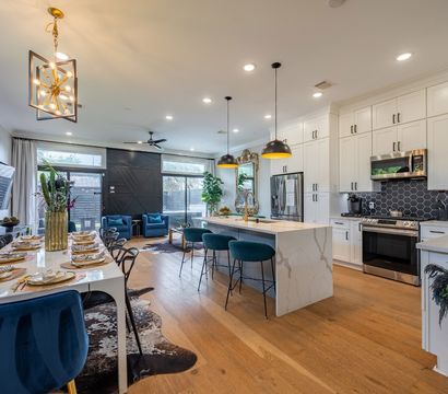 Contemporary open-plan kitchen and dining area with white shaker cabinets, marble waterfall island, black hex tile backsplash, brass pendant lights, teal bar stools, velvet dining chairs, hardwood floors and large glass sliding door.