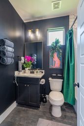 Compact modern bathroom with dark navy walls, white vanity and marble countertop, stacked gray towels, bright pink orchids, green velvet shower curtain, framed pink cat art and trailing potted plant by a small window