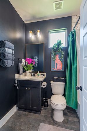 Compact modern bathroom with dark navy walls, white vanity and marble countertop, stacked gray towels, bright pink orchids, green velvet shower curtain, framed pink cat art and trailing potted plant by a small window