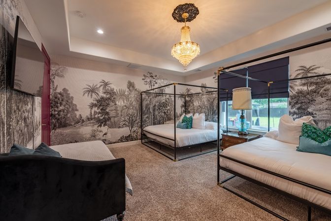Elegant bedroom with two black four-poster canopy beds, tropical black-and-white mural wallpaper, crystal chandelier, turquoise accent pillows, glass lamp on a wooden nightstand and plush carpet with a window view of a grassy backyard.