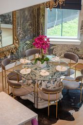 Stylish home dining room with octagonal glass-top table on an ornate white carved base, gray velvet chairs with gold frames, bright magenta orchid centerpiece, botanical wallpaper and a gilded mirror