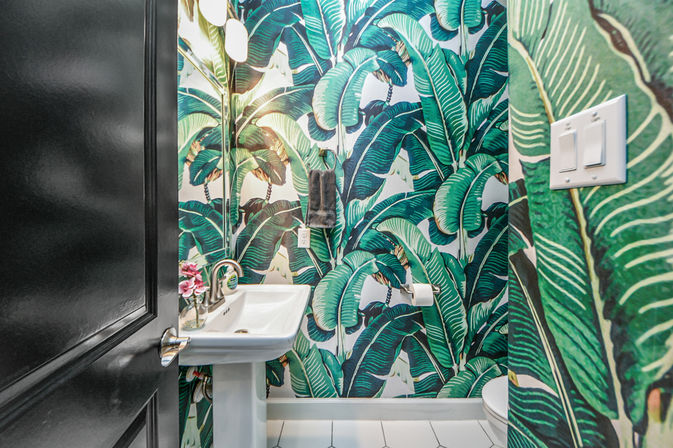 Tropical powder room with lush green banana-leaf wallpaper, white pedestal sink with brass faucet, small mirror and wall sconce, towel and toilet paper holder, and a dark entry door