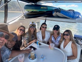Seven friends smiling around a table on a motorboat, enjoying drinks and sunny weather with inflatables stowed overhead and a forested shoreline in the background.