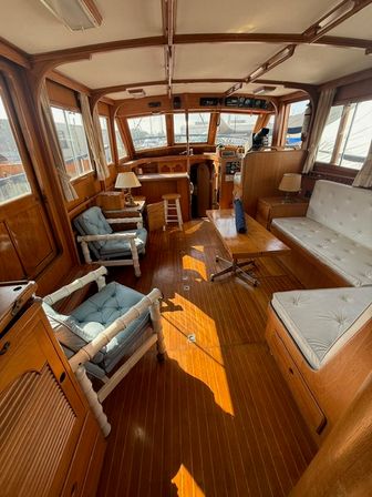 Sunlit wooden yacht salon with teak floor, cozy white tufted settee, blue cushioned armchairs, wooden coffee table and helm area overlooking a marina through large windows.