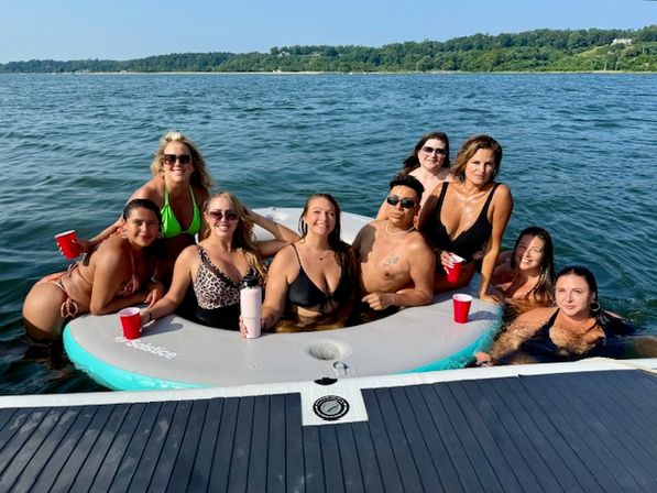 Group of friends relaxing on a circular inflatable float next to a boat dock on a sunny lake, wearing swimsuits and holding red cups for a summer boating vibe