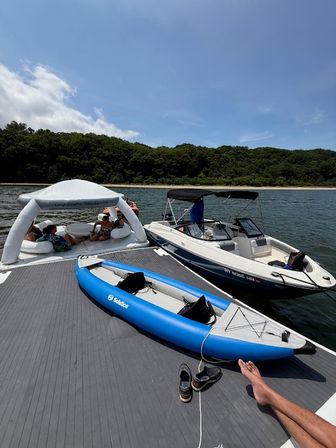 Sunny lake day: blue inflatable kayak tied to a floating dock, white motorboat moored nearby and people lounging under a white inflatable canopy with a tree-lined shoreline and clear blue sky.