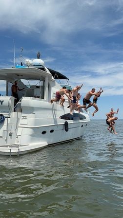 People jumping off a white yacht into calm coastal water under a bright blue sky — lively summer boat party scene