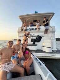 Eight friends in swimsuits posing between an inflatable dinghy and a white motor yacht at sunset on calm water — fun summer boating scene.