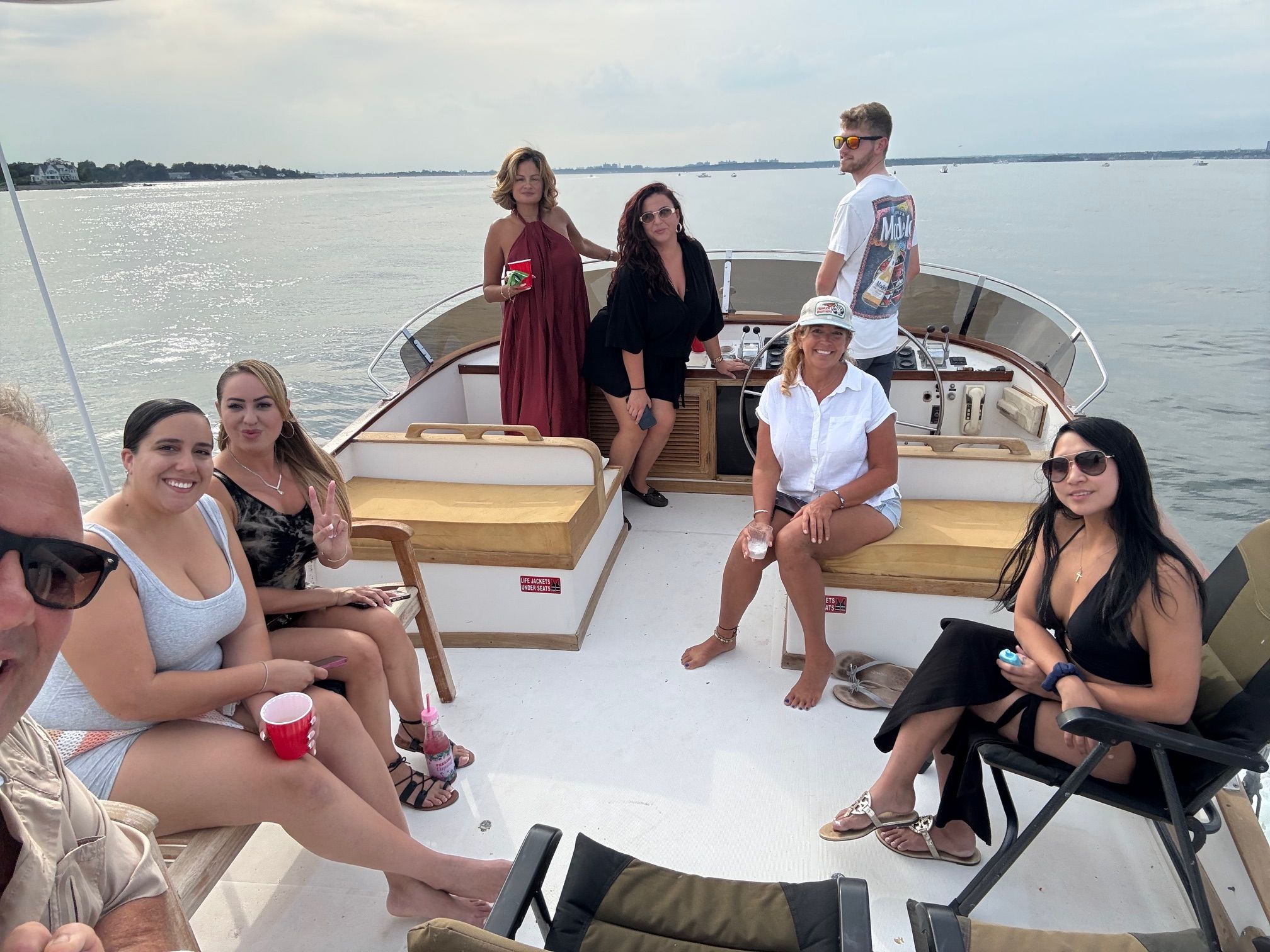 Group of eight friends relaxing on a motorboat deck in calm coastal waters, wearing summer clothes and sunglasses, sipping drinks and enjoying a daytime boat outing with shoreline in the distance.