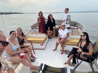Group of eight friends relaxing on a motorboat deck in calm coastal waters, wearing summer clothes and sunglasses, sipping drinks and enjoying a daytime boat outing with shoreline in the distance.