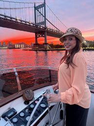 Smiling woman in a leopard-print hat steers a small motorboat under a lit suspension bridge at a vivid red-orange sunset, city skyline and glowing water reflections in the background.