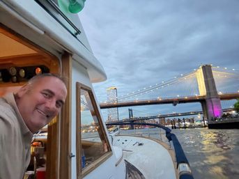 Cheerful person leaning from a small yacht cabin as the boat passes under the illuminated Brooklyn Bridge on the East River at dusk, Manhattan skyline and bridges lit in the background.