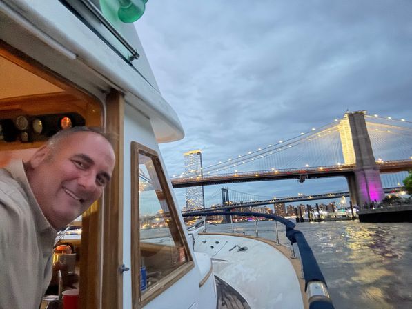 Cheerful person leaning from a small yacht cabin as the boat passes under the illuminated Brooklyn Bridge on the East River at dusk, Manhattan skyline and bridges lit in the background.