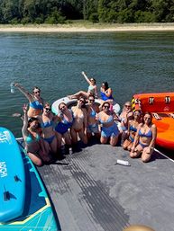 Cheerful group of women in colorful bikinis posing on a floating dock and inflatable tube at a sunny lake, paddleboard and forested shoreline in the background - summer lake party scene.
