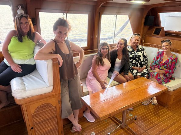 Six women smiling and relaxing in a teak-finished yacht cabin with white cushioned seating and a wooden table, sunlight reflecting on calm water through the windows — friends enjoying a sunset boat cruise.