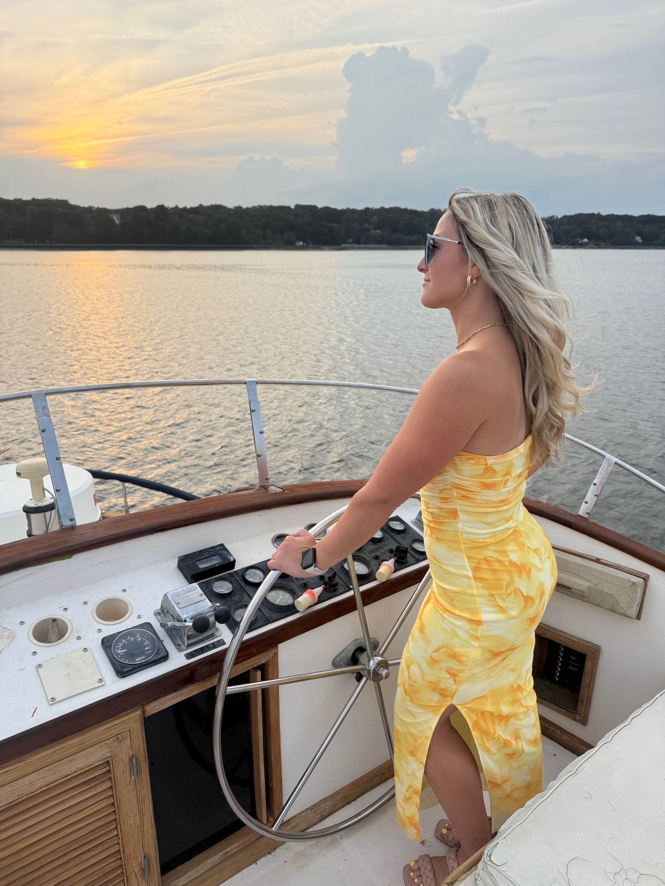 Person in a yellow floral dress steering a boat at sunset on a calm lake, sunglasses and windswept blonde hair with a tree-lined shoreline in the distance.