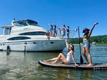 Group of women enjoying a sunny summer day on a white yacht and on a paddleboard in calm water, with blue sky and green shoreline in the background.