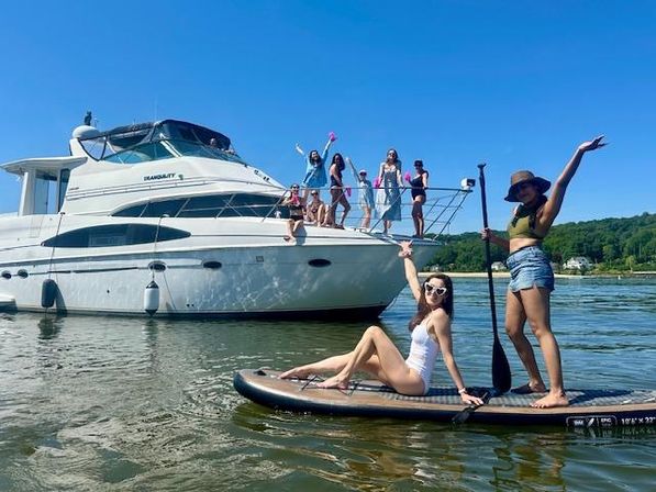 Group of women enjoying a sunny summer day on a white yacht and on a paddleboard in calm water, with blue sky and green shoreline in the background.