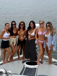 Six women in swimsuits smiling and holding drinks on a boat deck with calm water behind them — summer boating outing.