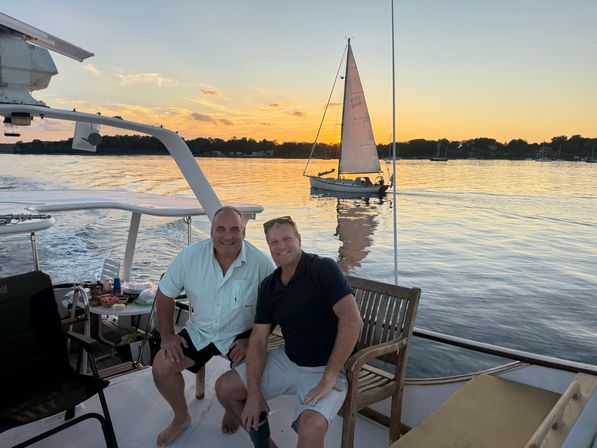 Two men smiling on a yacht at golden sunset, sailboat gliding on calm harbor waters with treeline silhouette.