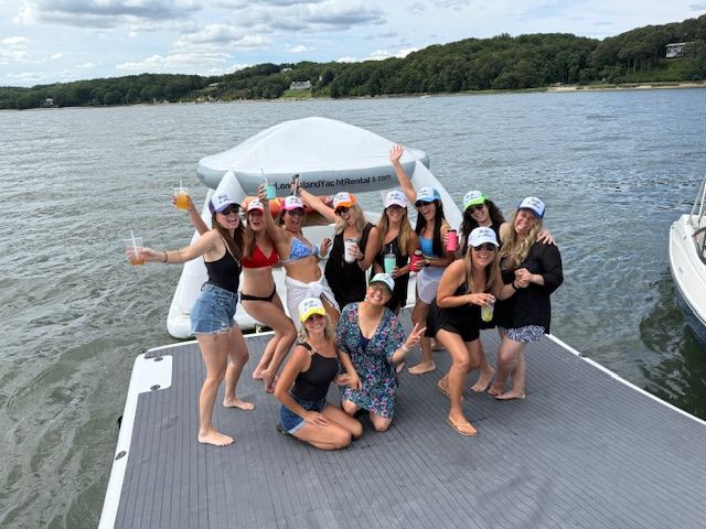Group of friends enjoying a summer boat party on a floating dock and inflatable lounge in a tree-lined bay, wearing swimsuits and caps while holding drinks and posing cheerfully.