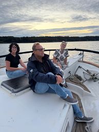 Three adults relaxing on a sailboat at sunset on a calm lake with a tree-lined shoreline and dramatic clouded sky