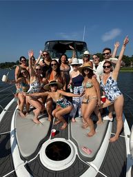Sunny summer boat party on a motorboat: a large group in swimsuits and sun hats posing and cheering on the bow with drinks, lake and tree-lined shoreline in the background.