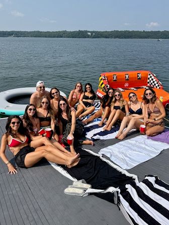 Group of friends lounging on a dock at a lake on a sunny summer day, wearing swimsuits and sunglasses, holding red cups beside inflatable floats and striped towels.