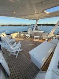 Yacht aft deck with teak flooring, white folding chairs and round table overlooking a calm coastal harbor with anchored boats and tree-lined shoreline at golden hour