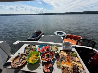 Summer boat picnic on a calm lake: a table stacked with corn on the cob, watermelon slices, salads, shrimp and a large charcuterie board, with a dinghy, inflatable tube and tree-lined shoreline in the background.