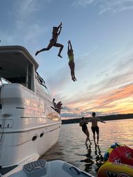 Two people in swimsuits mid-air jumping off a white yacht into a lake at sunset, onlookers on the deck, colorful sky reflected on calm water, inflatable towable and dinghy in the foreground.