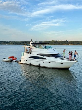 White motor yacht anchored on a calm lake at golden hour, friends on the bow with a dinghy and inflatable tube trailing, wooded shoreline in the background