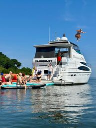 People on paddleboards gathered around a white yacht in a calm blue bay under a clear sky, one person midair jumping from the top deck.