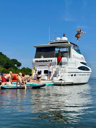 People on paddleboards gathered around a white yacht in a calm blue bay under a clear sky, one person midair jumping from the top deck.