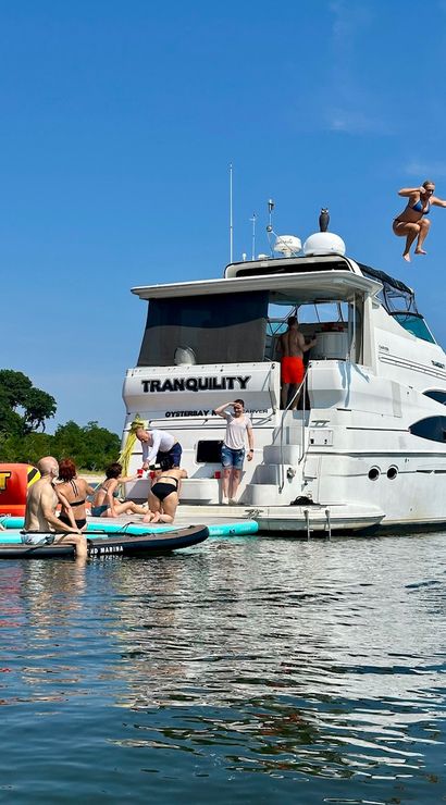 People on paddleboards gathered around a white yacht in a calm blue bay under a clear sky, one person midair jumping from the top deck.