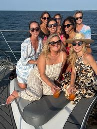 Eight women in sunglasses and summer dresses laughing together on a yacht deck, holding cocktails with the open ocean and distant coastline behind them.