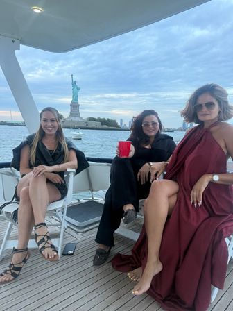 Three women relaxing on a yacht near the Statue of Liberty in New York Harbor at dusk, one holding a red cup, wearing summer outfits and sunglasses, wooden deck and cloudy skyline