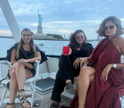 Three women relaxing on a yacht near the Statue of Liberty in New York Harbor at dusk, one holding a red cup, wearing summer outfits and sunglasses, wooden deck and cloudy skyline