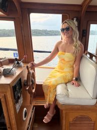 Smiling woman in a yellow sundress and sunglasses at a wooden boat helm inside a cabin, steering wheel and controls visible with a calm lake and tree-lined shore outside the windows — relaxed summer boat cruise scene.