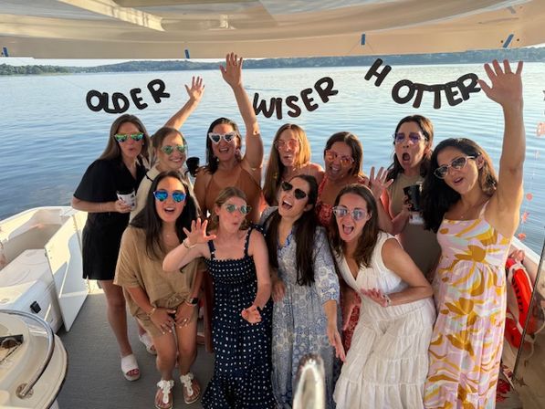 Group of women in sunglasses and summer dresses celebrating on a pontoon boat on a lake under a playful "Older Wiser Hotter" banner