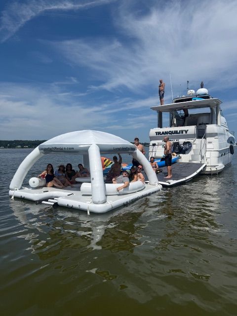 Summer boat party on a calm lake — group lounging on a white inflatable floating island tethered to a yacht under a bright blue sky with wispy clouds.