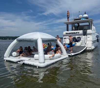 Summer boat party on a calm lake — group lounging on a white inflatable floating island tethered to a yacht under a bright blue sky with wispy clouds.