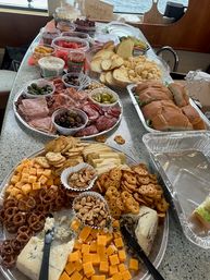 Party-ready spread on a boat counter: charcuterie and cheese platters with cubed cheddar, blue cheese, pretzels, crackers, nuts, olives, sliced baguette sandwiches, chips and fruit with water view.