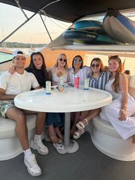 Group of six friends smiling around a white table on a yacht at sunset, canned drinks on the table, inflatable paddleboards stowed overhead and sailboats on a lake in the background