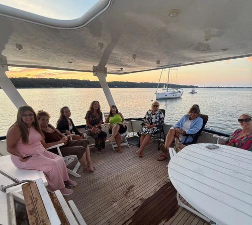 Eight women relaxing on a covered boat deck at sunset, wooden floor and round table visible, with a sailboat anchored on calm lake waters in the background.