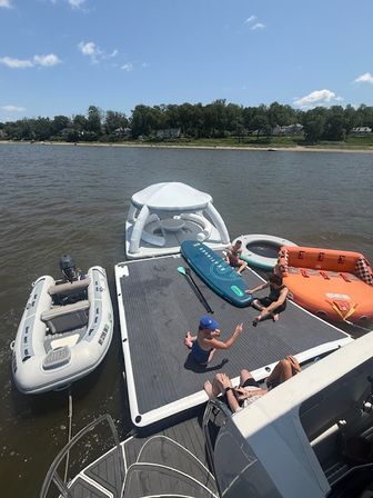 Sunny river scene: people relaxing on a boat’s large swim platform with an inflatable dinghy, white floating lounge, paddleboard and orange towable tube, tree-lined shoreline and blue sky in the background.